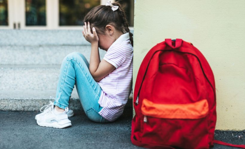 young girl with back up against wall in school yard with red backpack on ground