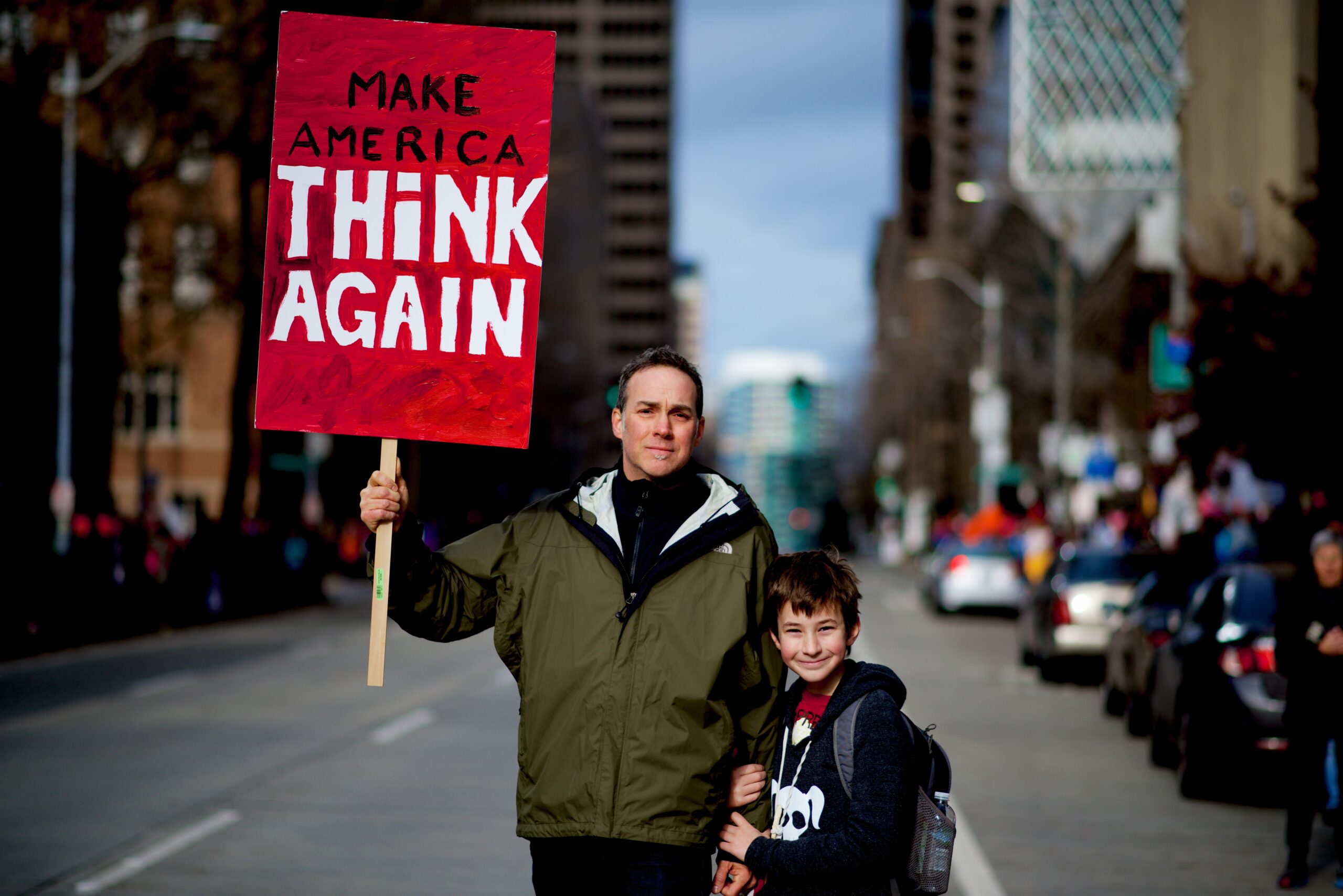 Child stands proudly with his father as his dad holds a sign that reads "Make America Think Again", setting a good example of how we need to stand up for what is right.