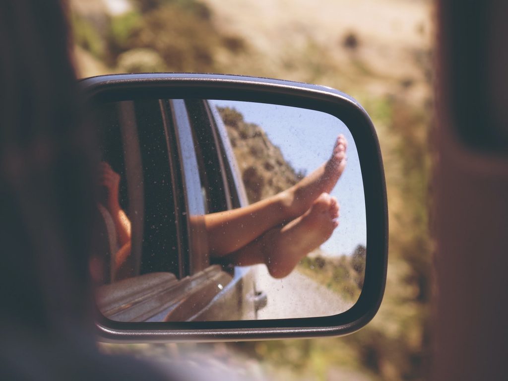 A car's side-view mirror reflects a pair of legs hanging out the backseat window, suggesting a road trip, a perfect thing to do with teenagers