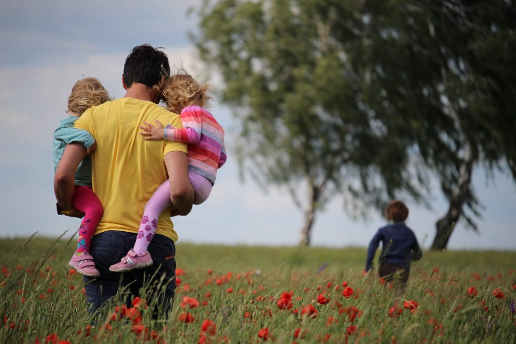 A father carries his toddlers—who look like they're in the terrible two age—across a field of flowers, following their older brother who's running ahead