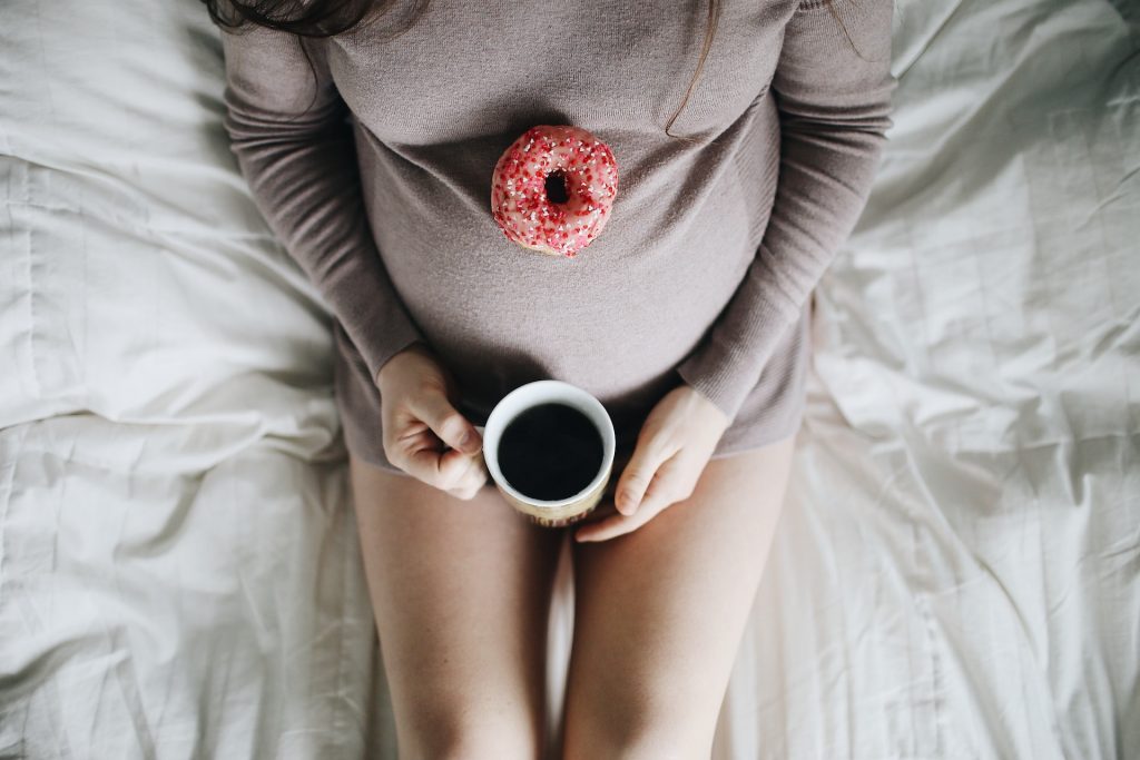 An expectant mom is shot from above. She's sitting on the bed, holding a cup of black coffee while a donut with sprinkles rests on top of her pregnant belly.
