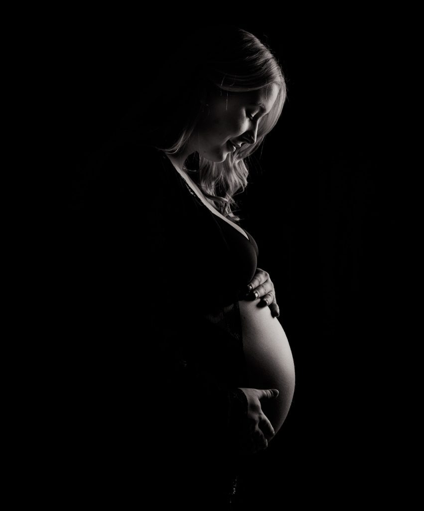 In a black-and-white photo, the light catches just a pregnant woman's belly and her facial features. The rest is left in darkness. 