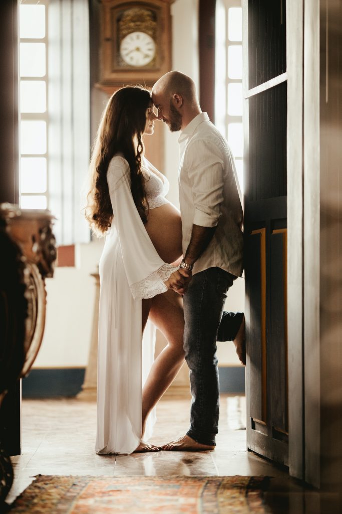Expectant parents hold hands and touch foreheads while daddy stands against a wall. 