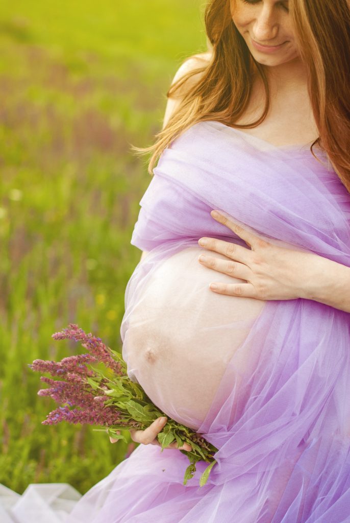 A pregnant woman in a wispy lavender dress stands in the middle of a field while holding lavender blooms