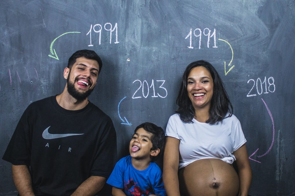 Parents and their pre-schooler pose against a chalkboard, smiling. Their birth years are printed on the board, including one for the baby in mom's tummy. 