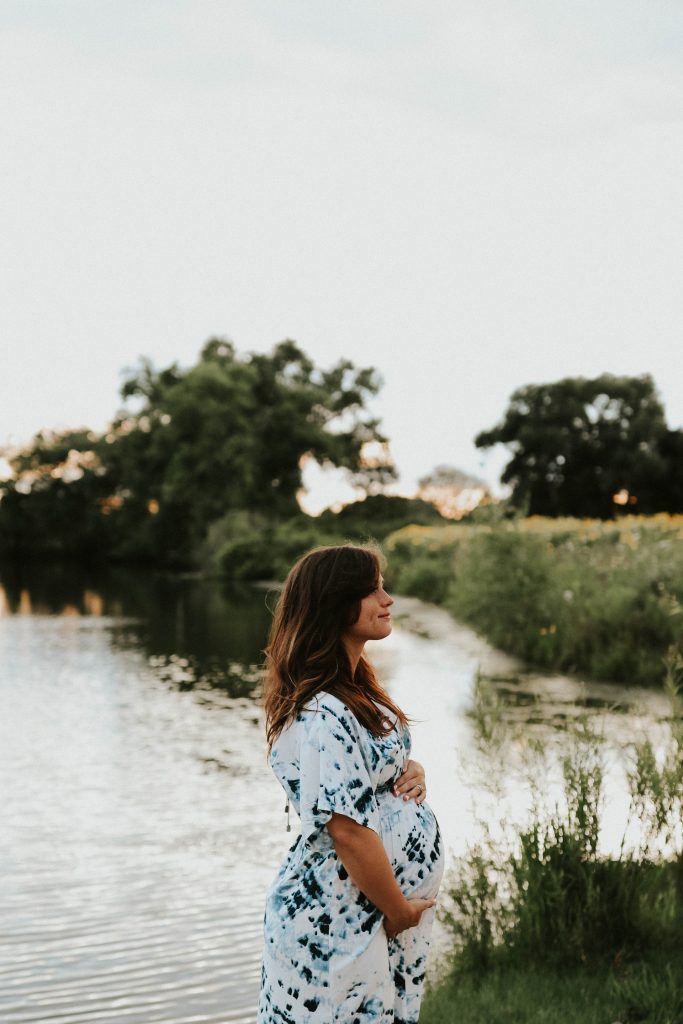 A woman cradles her baby bump with both hands while standing by a lake, looking away from the camera