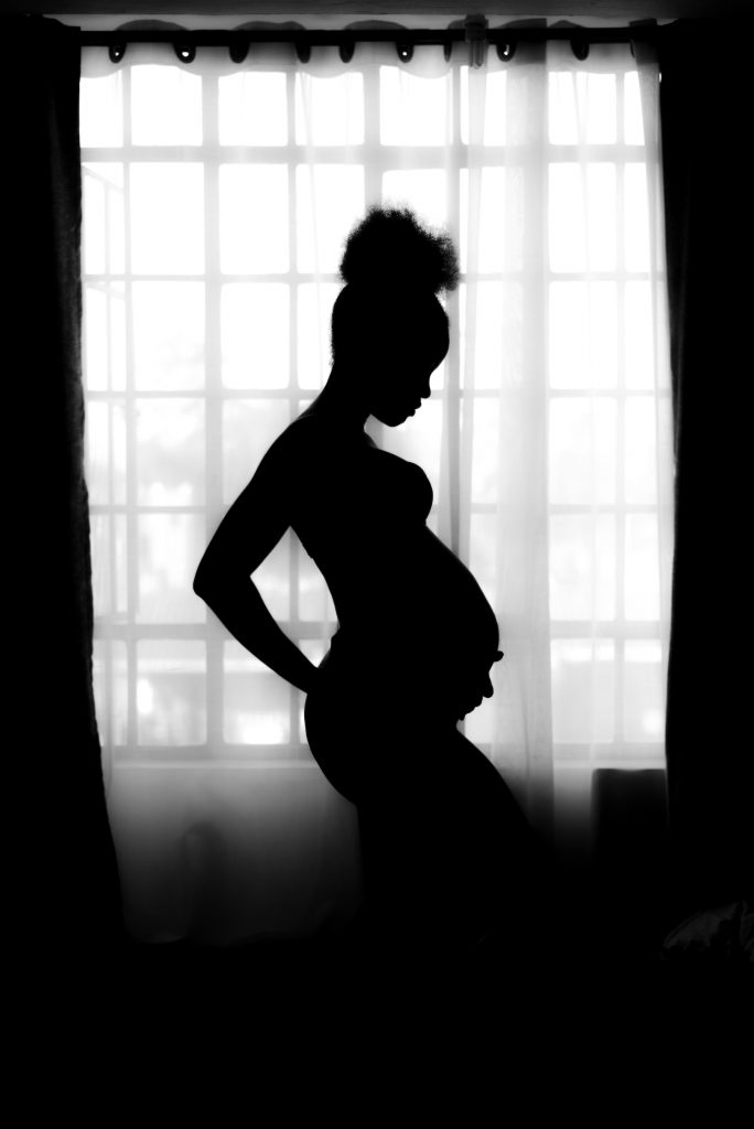 The silhouette of expectant mom in front of a window with the drapes open. 