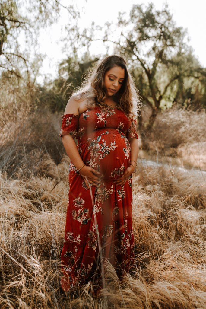 An expectant mom wearing a red floral dress stands in the middle of an overgrown park. 