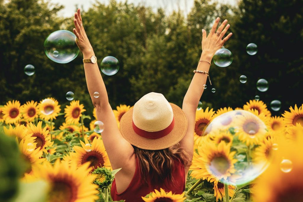 A woman, with her back to the camera, raises her arms with glee as she stands in the middle of a sunflower farm, with bubbles floating around her. 