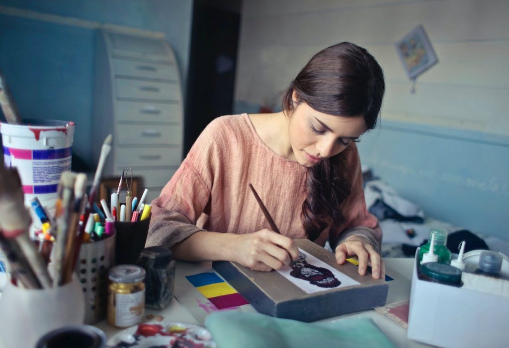 A woman sits at a desk in her studio—which is full of art equipment—painting. 