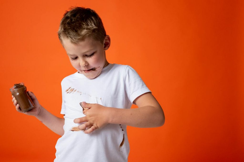 A boy, with chocolate spread on his face, smears the same spread on his white t-shirt.
