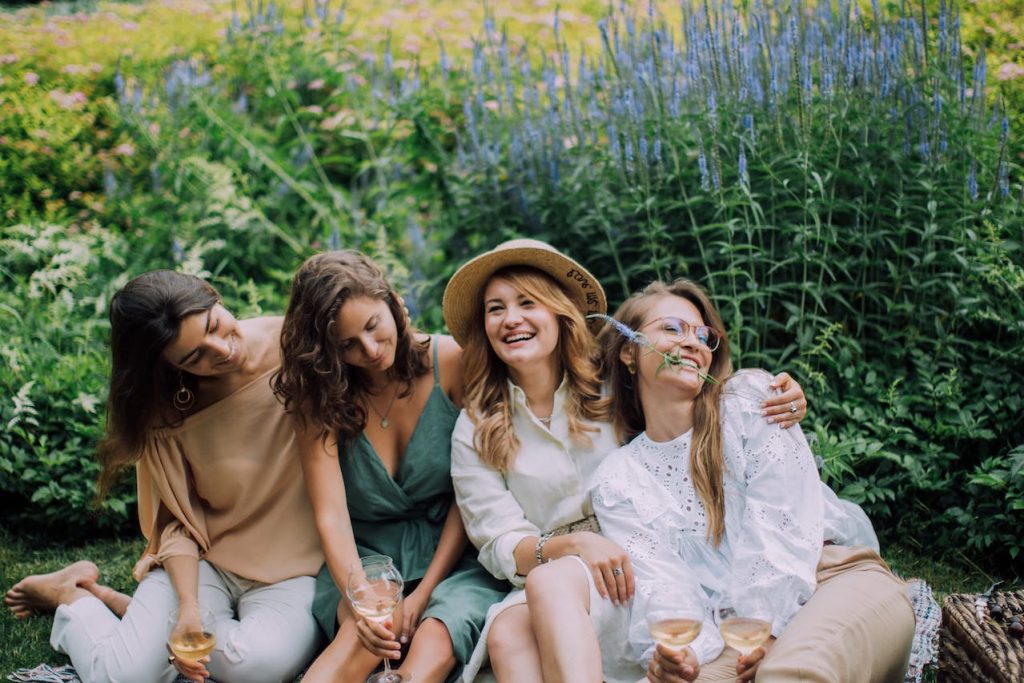 Four women sit and laugh together on the grass with lavender plants behind them. 