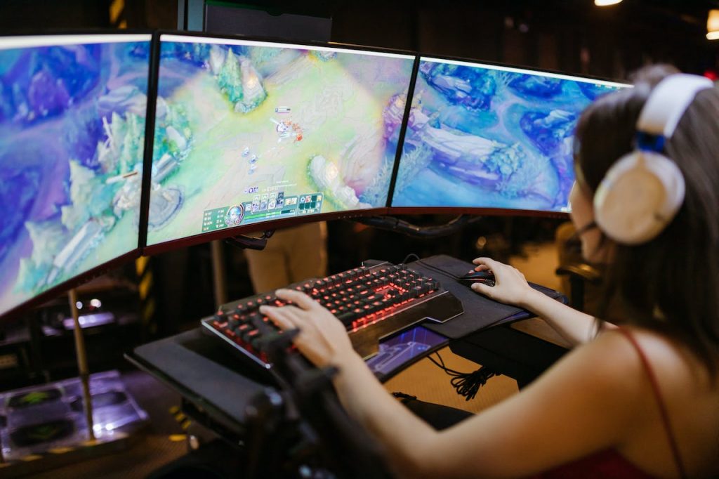 A woman wearing headphones plays a video game across three monitors. 