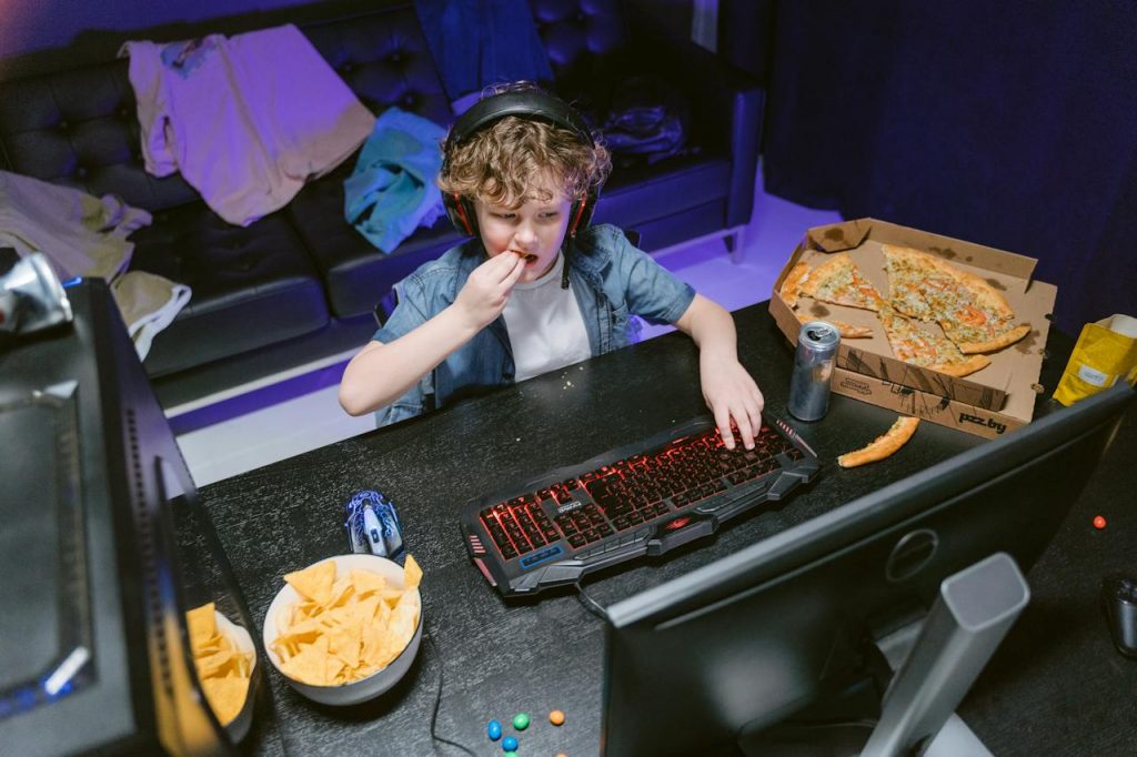 A pre-teen boy wears headphones while sitting at a desk in front of a gaming computer and eating pizza and chips. 