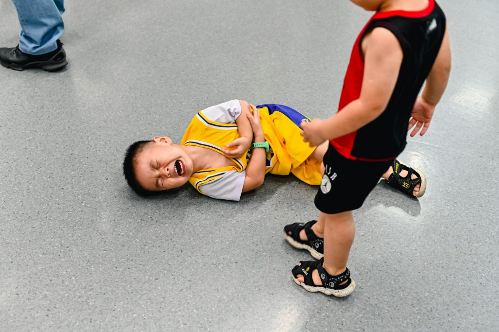 A boy lies on the floor, crying in pain while clutching his elbow while another boy stands over him. 