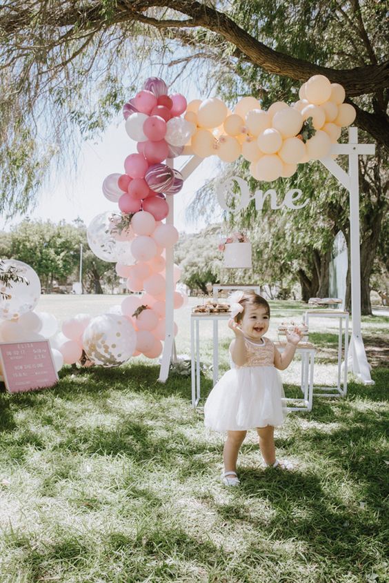 Smiling little girl in front of an outdoor birthday party setup with lots of balloons.