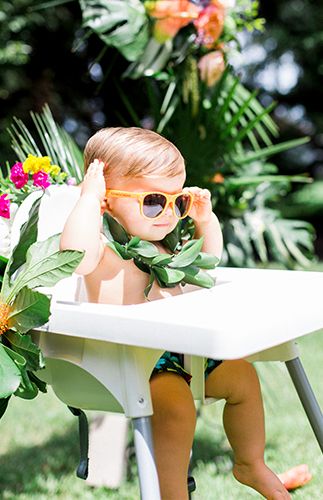 Baby boy on a high chair wearing leis and sunglasses.