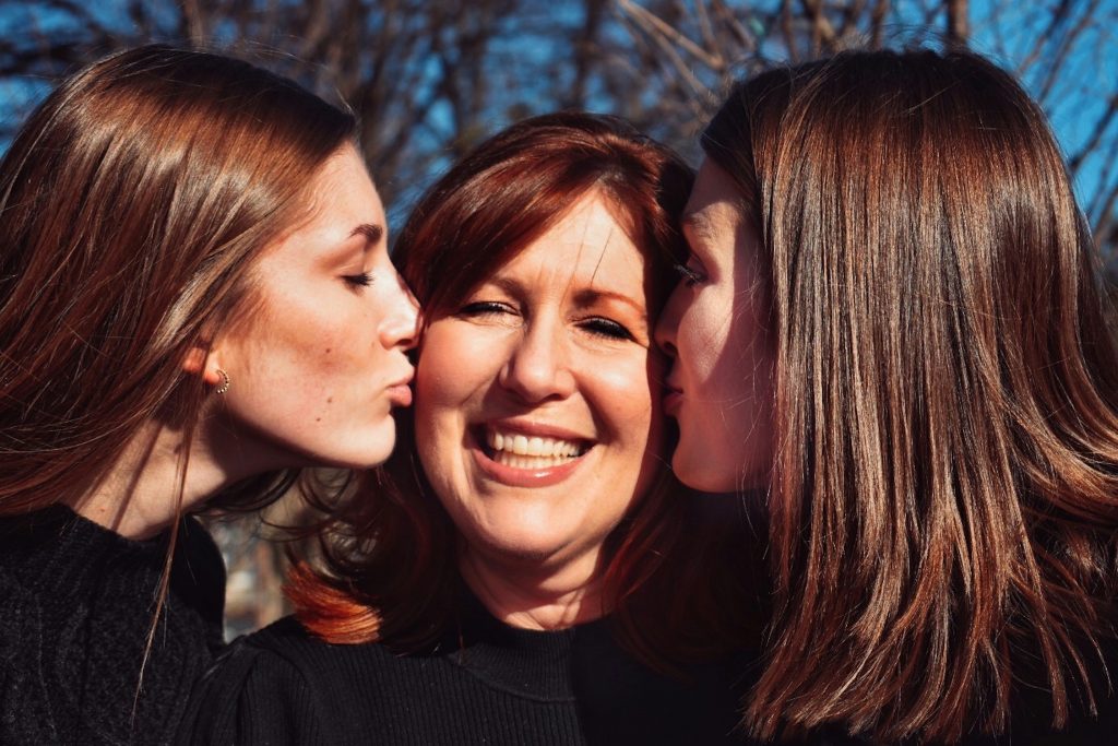 Smiling woman while being kissed by daughters on her cheeks.