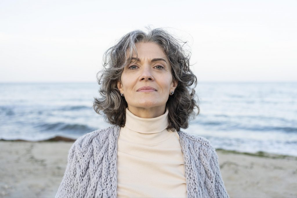 Older woman with grey curly hair on a beach looking straight at the camere.