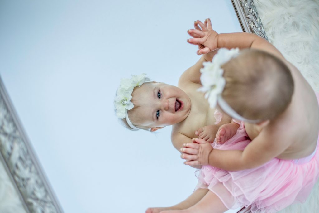 Baby girl smiling at a mirror while sitting down.