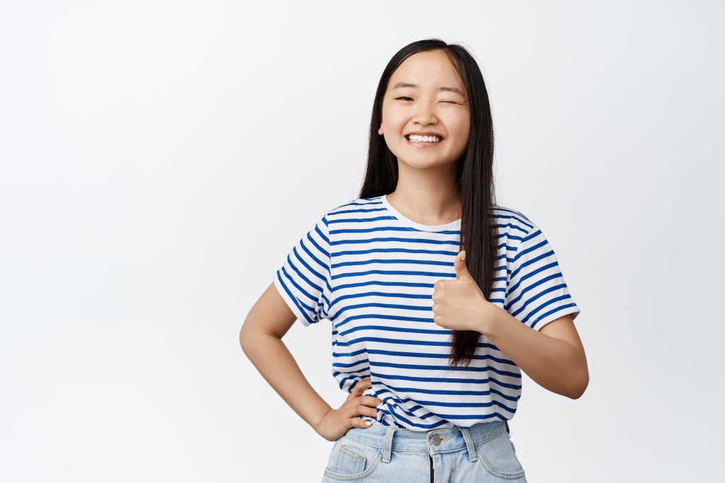 Smiling Asian teen and winking at the camera while showing thumbs up.