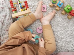 A close up picture of a child from the tummy down as they sit on the floor wearing brown overalls and playing eith stacking toys.
