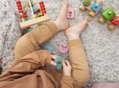A close up picture of a child from the tummy down as they sit on the floor wearing brown overalls and playing eith stacking toys.